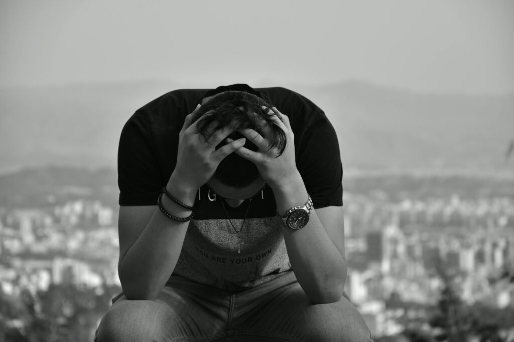 A focused young man sits with hands on head against an urban cityscape in grayscale, suggesting introspection. Pecados sins